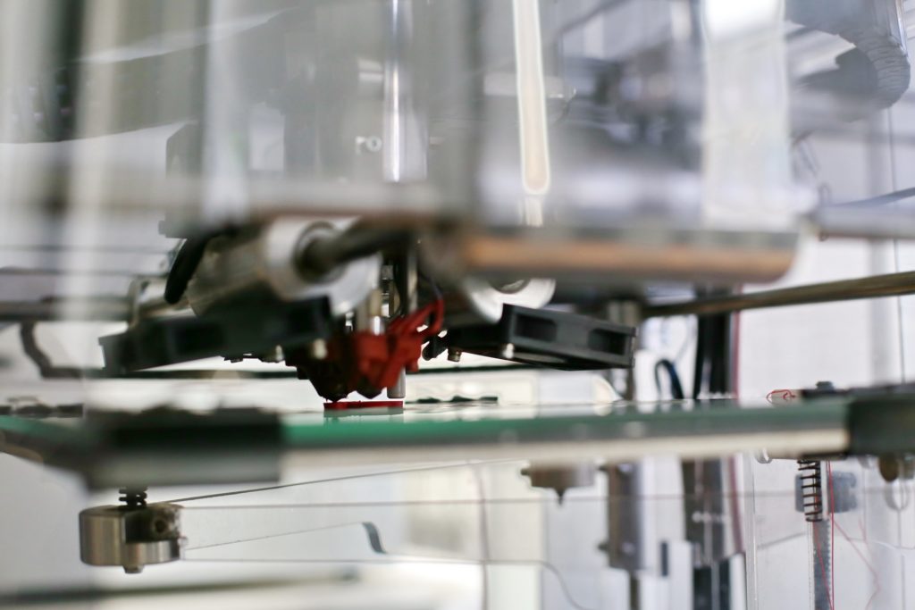 Drukarka 3D drukuje czerwony element na zielonej platformie. Widoczne są mechaniczne części drukarki.