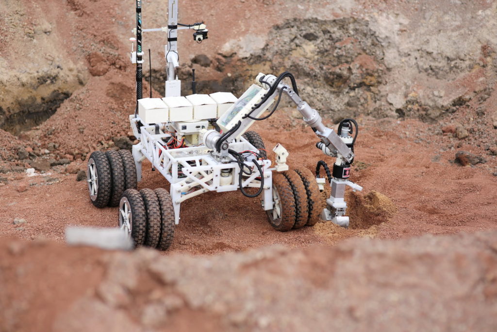 Robot z ramieniem i kamerą na kołach, badający teren. Kontekst: eksploracja.