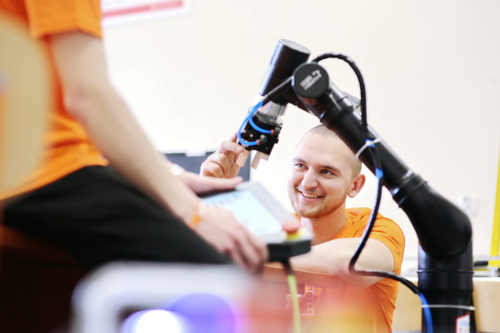 Mężczyzna w pomarańczowej koszulce pracuje z robotem. W tle widać tablet.