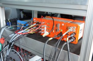 Pomarańczowe i niebieskie moduły elektroniczne z licznymi kablami. Kontekst: sprzęt pomiarowy w pojeździe.