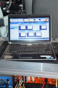 Laptop z oprogramowaniem diagnostycznym, podłączony do urządzeń z kablami. Kontekst: wnętrze pojazdu, warsztat.