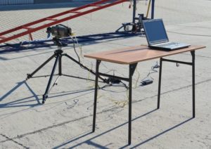 Kamera na statywie i laptop na stole. Sprzęt ustawiony na zewnątrz, w pobliżu konstrukcji.