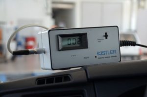 Urządzenie Kistler z wyświetlaczem cyfrowym, podłączone kablem. Znajduje się na desce rozdzielczej pojazdu.