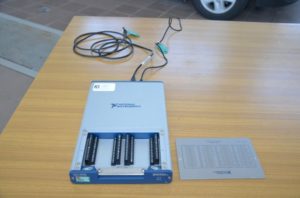 Urządzenie National Instruments z gniazdami i kablami. Obok leży arkusz z danymi.