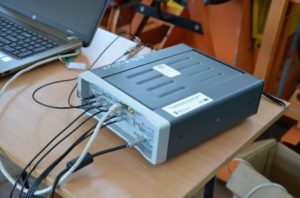 Urządzenie pomiarowe z wieloma złączami i kablem podłączonym do laptopa. Kontekst: laboratorium lub warsztat.