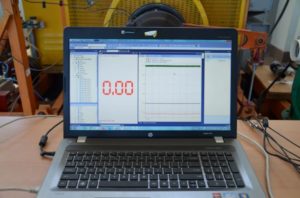 Laptop z ekranem pokazującym odczyt 0.00 i wykres. W tle maszyna.