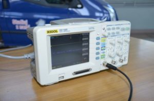 Oscyloskop cyfrowy Rigol DS1102D z podłączonym kablem. W tle niebieski pojazd.