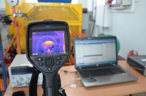 Kamera termowizyjna FLIR E53 i laptop HP na stole. W tle żółta maszyna. Kontekst: testy, pomiary.