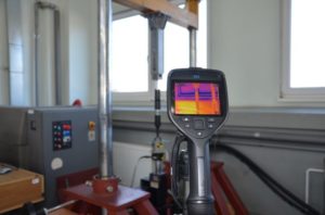 Kamera termowizyjna pokazuje rozkład temperatury na elemencie maszyny. Kluczowe obiekty: kamera termowizyjna, maszyna, okno.