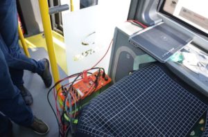 Elektronika i laptop na siedzeniu w pojeździe. W tle widoczne nogi pasażerów i żółte poręcze.