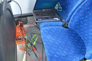 Laptop i pomarańczowa skrzynka z przewodami na siedzeniu autobusu. Kontekst: diagnostyka lub testowanie sprzętu.