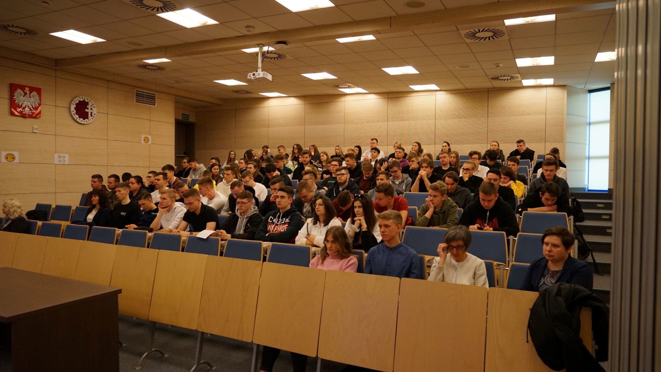 Sala wykładowa z grupą studentów siedzących w ławkach. W tle widoczne godło Polski i logo uczelni.