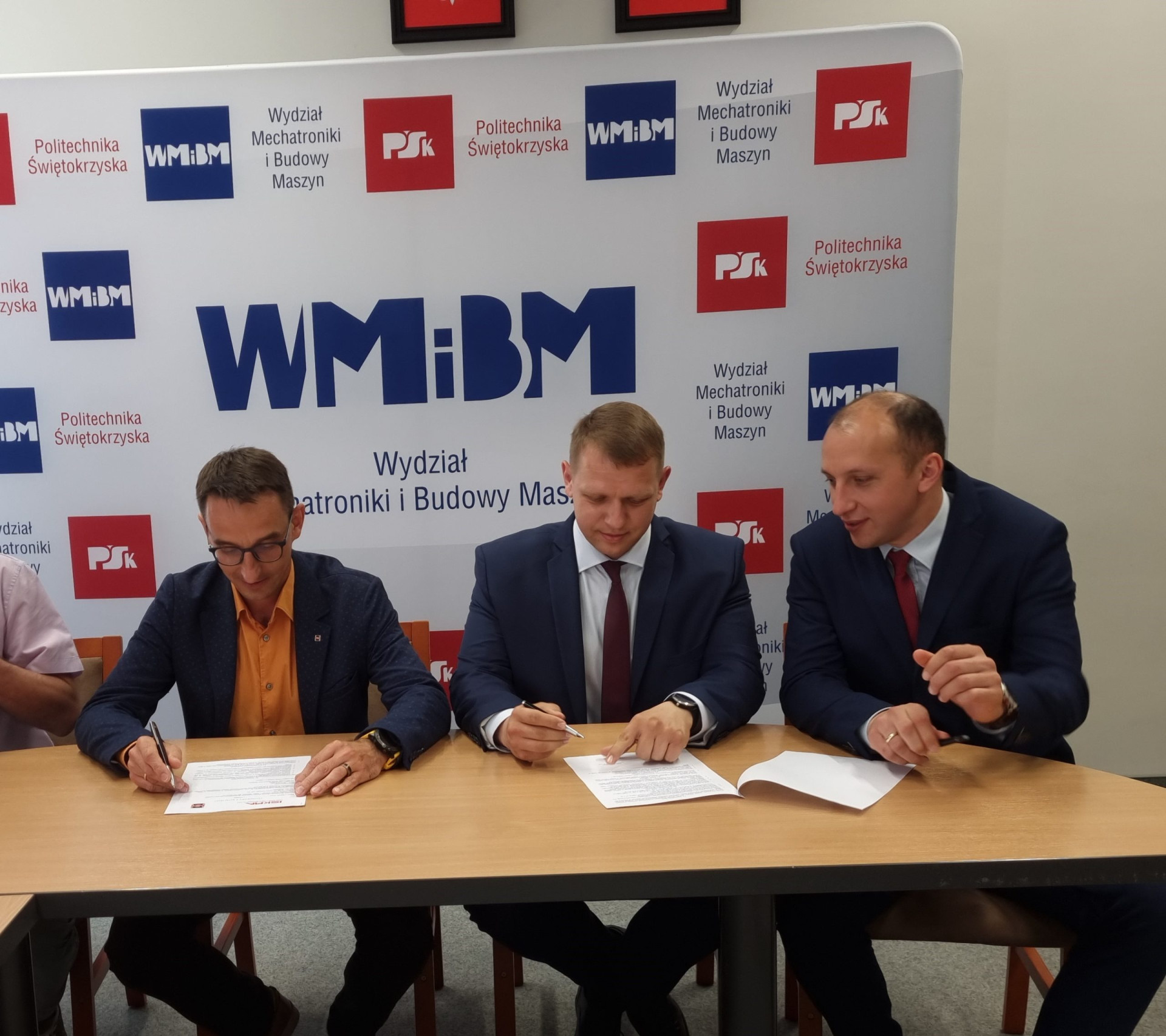 Trzech mężczyzn w garniturach podpisuje dokumenty przy stole. W tle logo Politechniki Świętokrzyskiej.