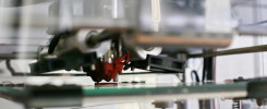 Drukarka 3D drukuje czerwony element na zielonej platformie. Widoczne są mechaniczne części drukarki.