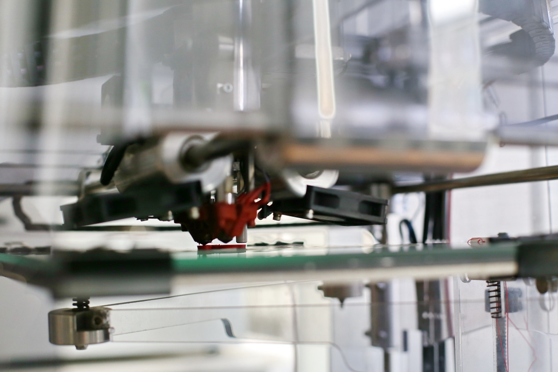 Drukarka 3D drukuje czerwony element na zielonej platformie. Widoczne są mechaniczne części drukarki.