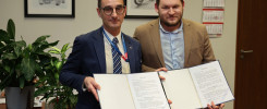 Dwóch mężczyzn trzyma otwarty dokument. W tle obrazy samochodów, motocykla i kalendarze.