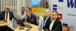 Mężczyźni siedzą przy stole, na którym znajdują się butelki z wodą, filiżanki i laptop. Kontekst to spotkanie lub konferencja.