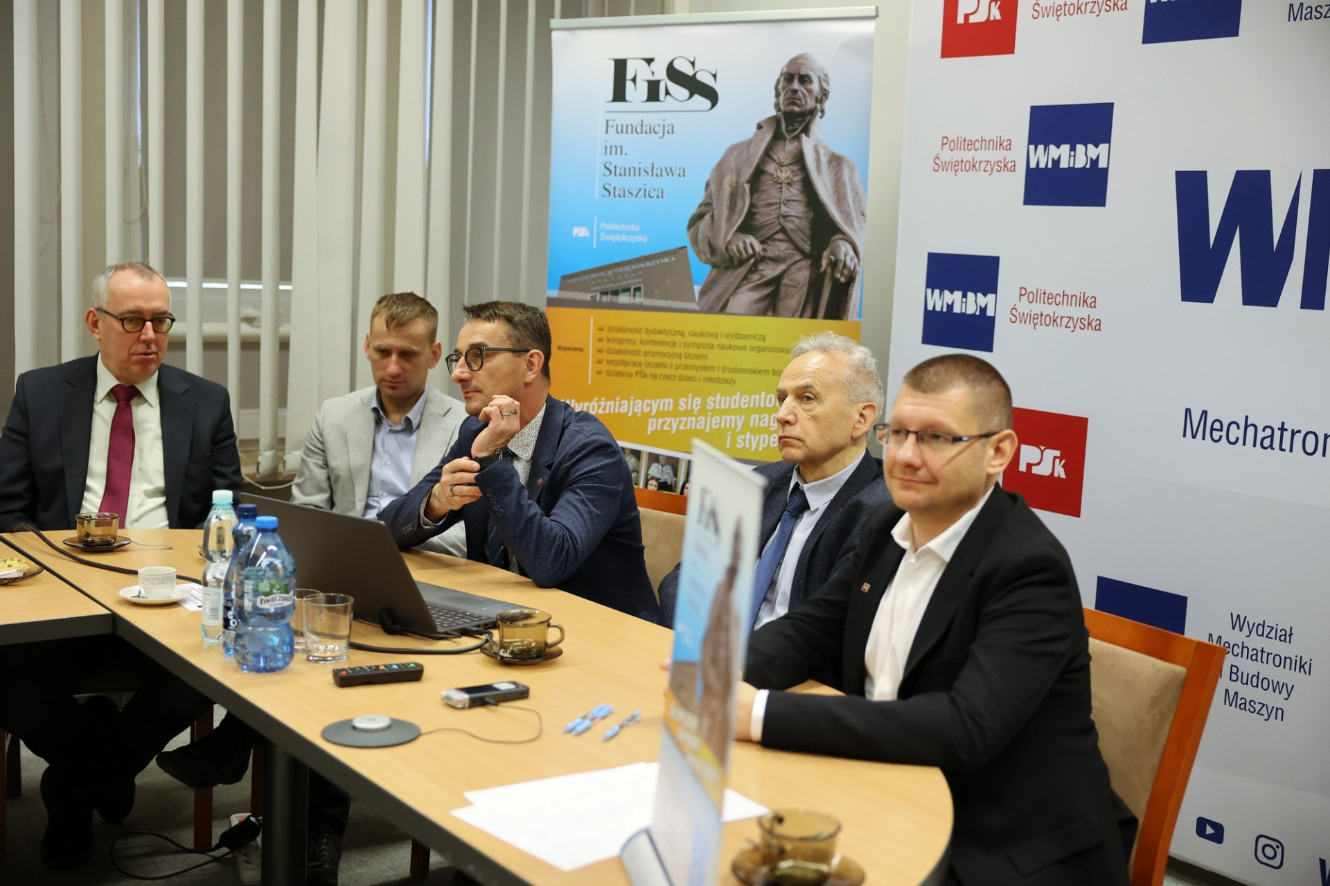 Mężczyźni siedzą przy stole, na którym znajdują się butelki z wodą, filiżanki i laptop. Kontekst to spotkanie lub konferencja.