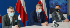 Trzech mężczyzn w garniturach siedzi przy stole. Na stole mikrofon. W tle flagi Polski i baner.