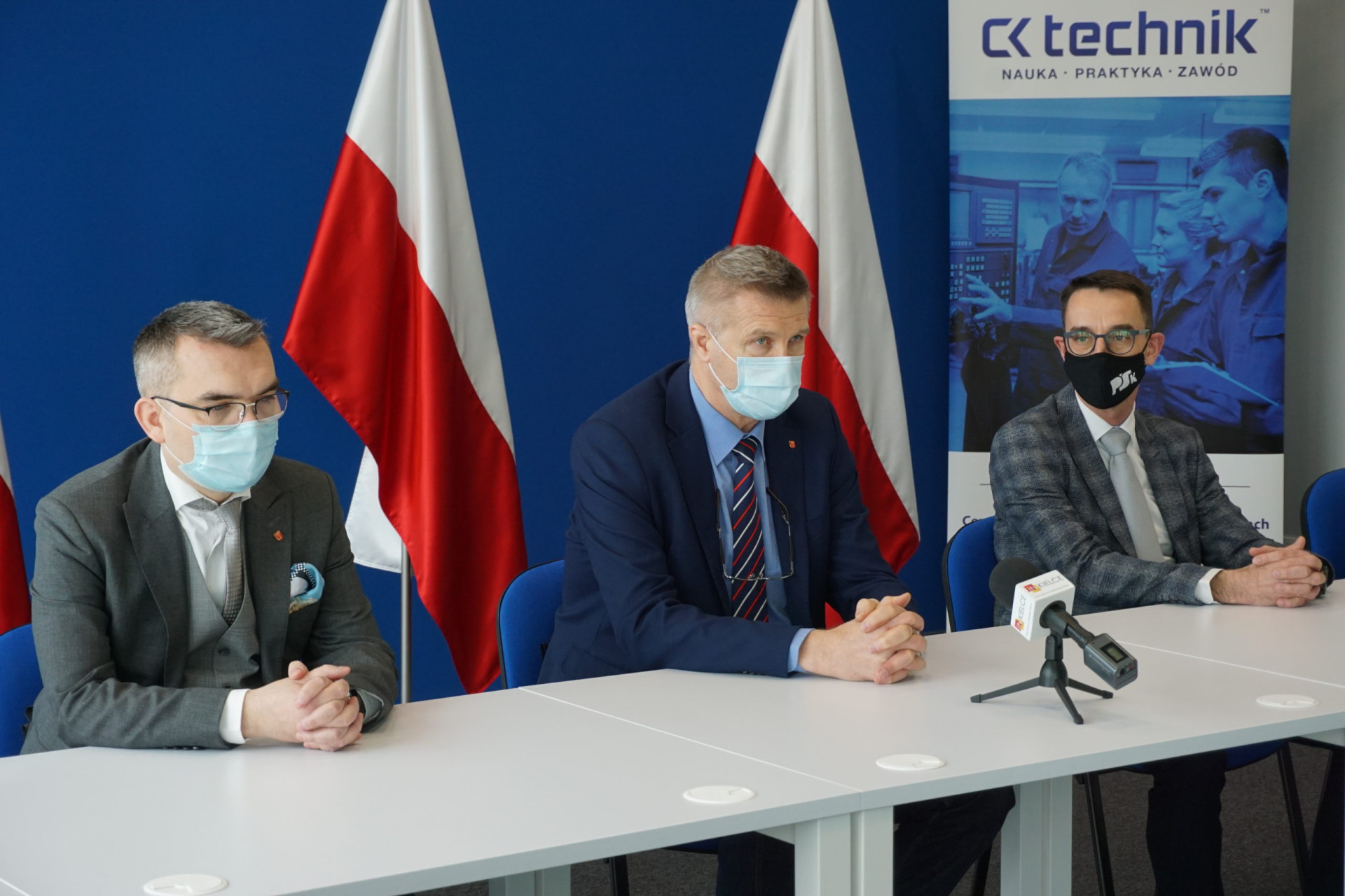Trzech mężczyzn w garniturach siedzi przy stole. Na stole mikrofon. W tle flagi Polski i baner.