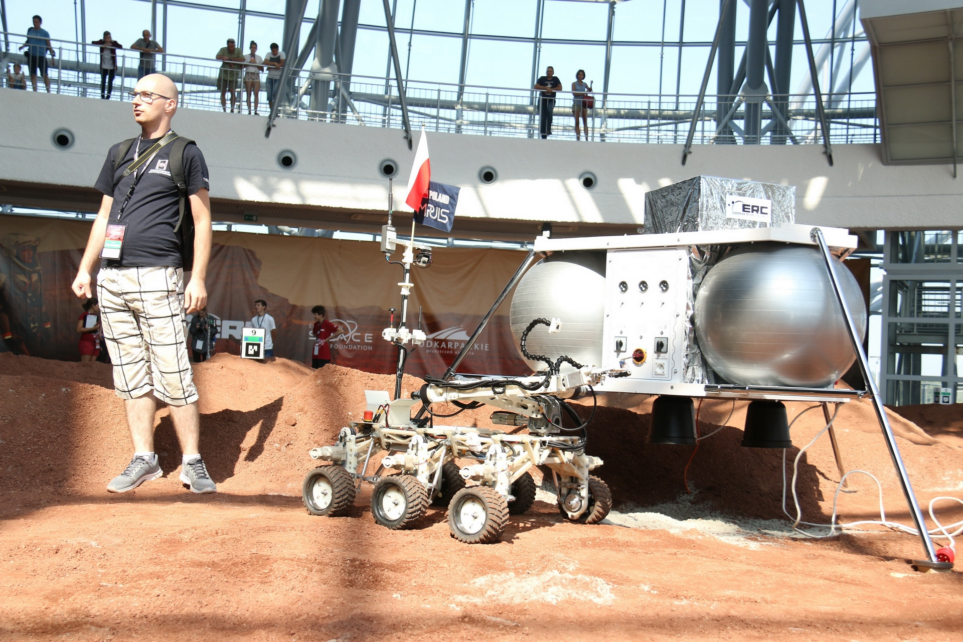 Robot marsjański z polską flagą. W tle ludzie i konstrukcja budynku.