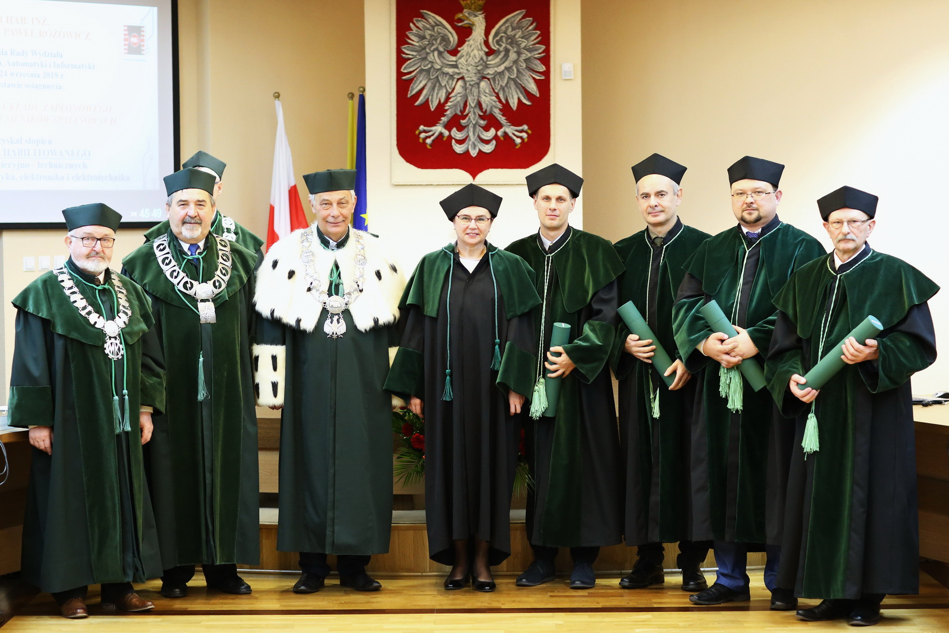 Grupa osób w togach akademickich pozuje do zdjęcia. Kluczowe obiekty: togi, czapki, dyplomy. Kontekst: uroczystość akademicka.