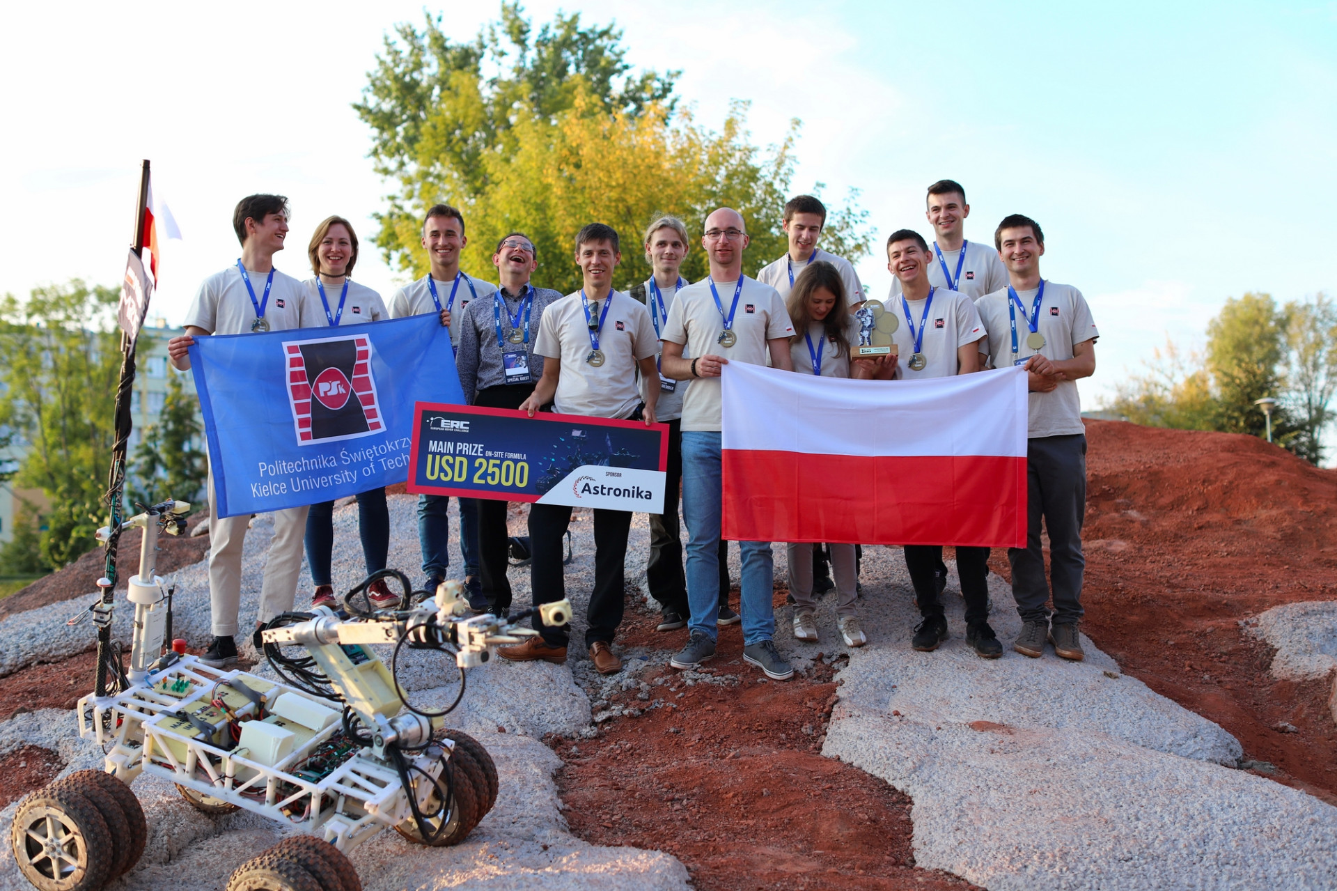 Grupa młodych ludzi z Politechniki Świętokrzyskiej z flagą Polski i nagrodą. Kluczowe obiekty: robot, flagi, nagroda, ludzie.