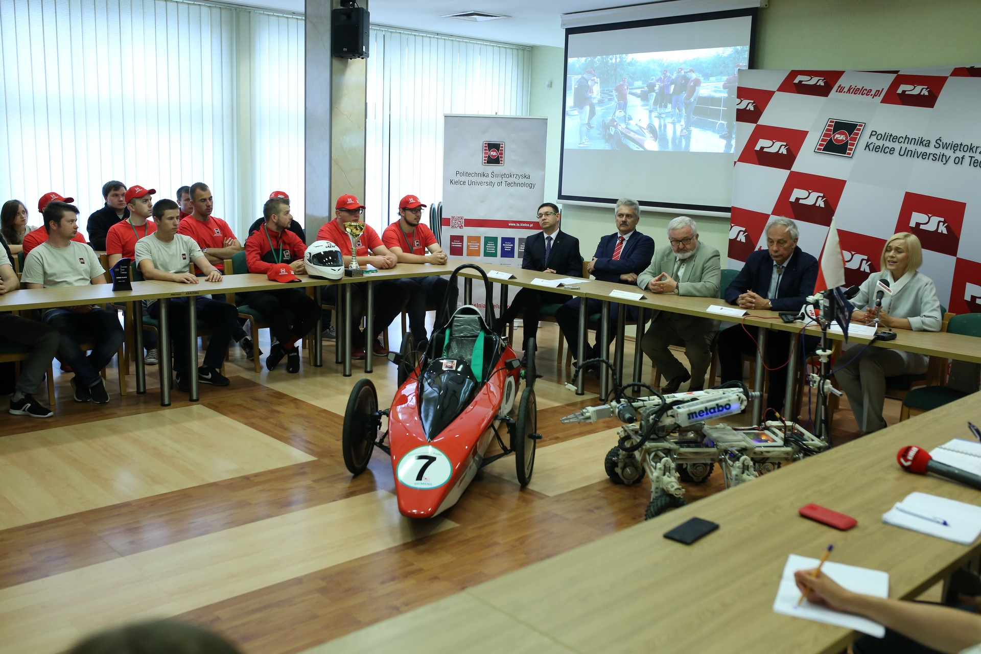 Grupa osób siedzi przy stole, przed nimi bolid wyścigowy i robot. Kontekst: wydarzenie na Politechnice Świętokrzyskiej.