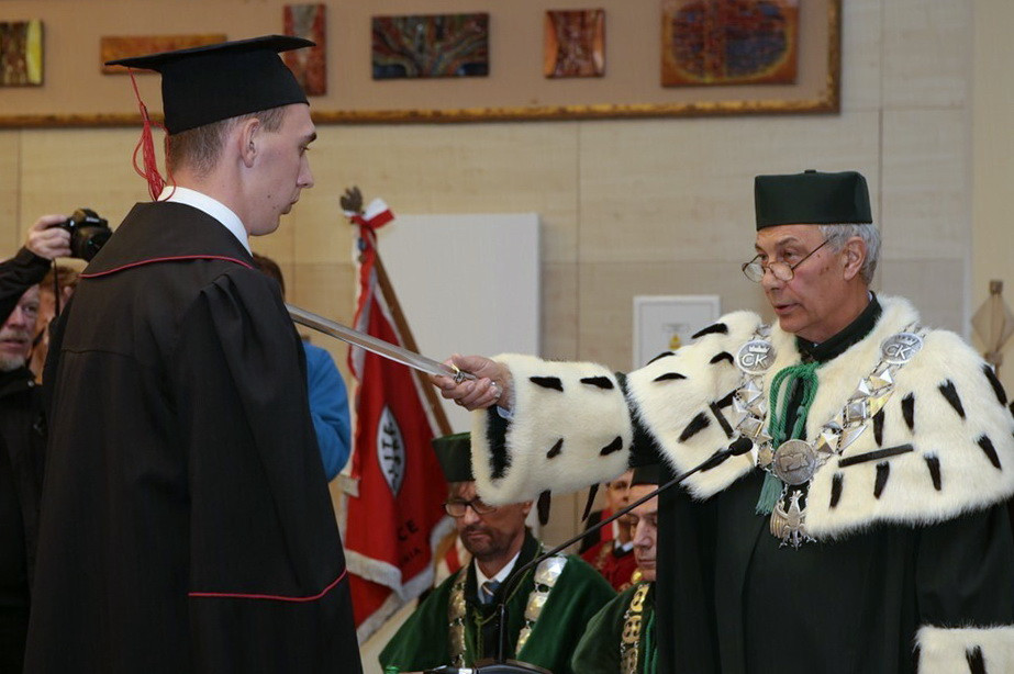 Uroczystość akademicka. Profesor w todze z futrem przekazuje miecz studentowi w czarnej todze i birecie.