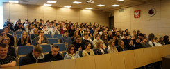 Grupa studentów siedzi na wykładzie. W tle widoczne godło Polski i logo uczelni.
