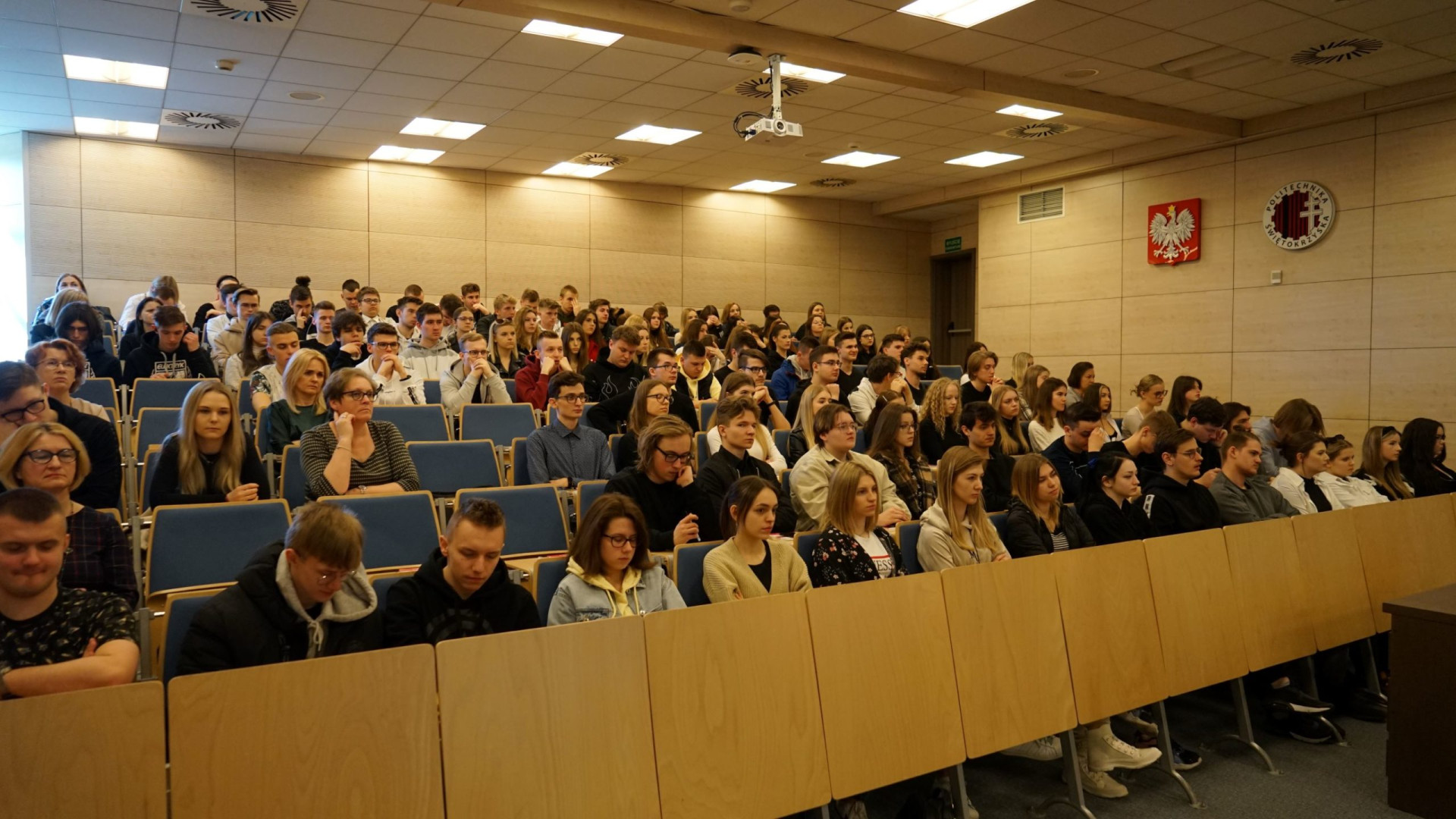 Grupa studentów siedzi na wykładzie. W tle widoczne godło Polski i logo uczelni.