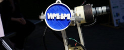 Prototypowy robot z niebieskim dyskiem z logo WMiBM. Obiekty: robot, dysk, logo. Kontekst: wystawa naukowa lub technologiczna.