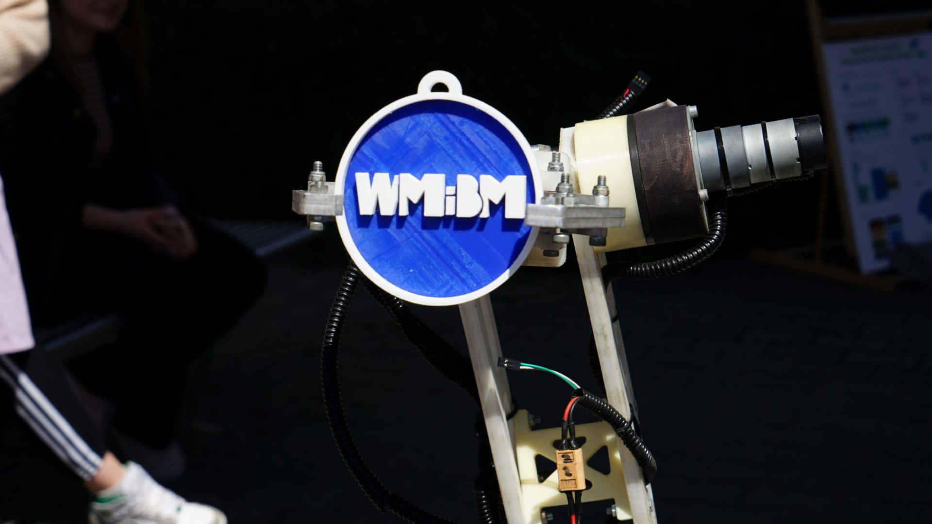Prototypowy robot z niebieskim dyskiem z logo WMiBM. Obiekty: robot, dysk, logo. Kontekst: wystawa naukowa lub technologiczna.