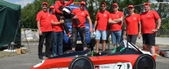 Grupa młodych ludzi w czerwonych koszulkach pozuje z pojazdem wyścigowym. Kluczowe obiekty: zespół, pojazd, kask.
