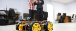 Zbudowany robot mobilny z żółtymi kołami i elektroniką. W tle dwie osoby w pomieszczeniu biurowym.