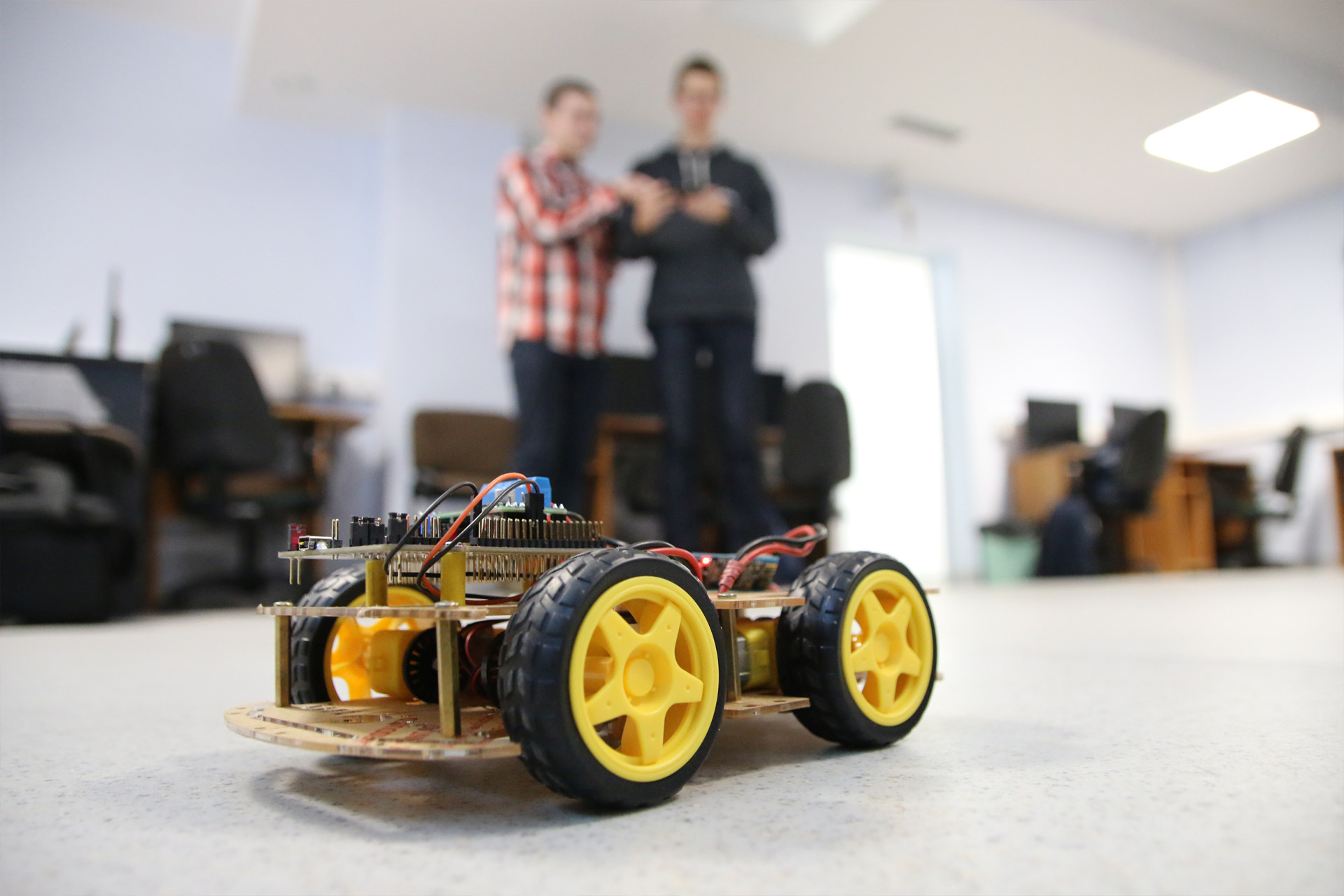 Zbudowany robot mobilny z żółtymi kołami i elektroniką. W tle dwie osoby w pomieszczeniu biurowym.