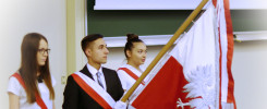 Trzy osoby w strojach galowych, z biało-czerwonymi szarfami, niosą polską flagę. Kontekst: uroczystość szkolna.