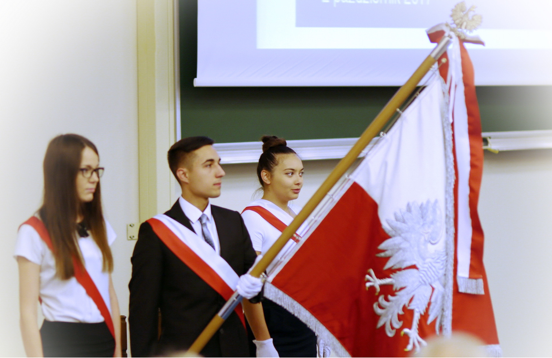 Trzy osoby w strojach galowych, z biało-czerwonymi szarfami, niosą polską flagę. Kontekst: uroczystość szkolna.