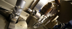Cylindryczny element z wycięciami w uchwycie maszyny. Obok głowica maszyny z dyszą. Kontekst: produkcja przemysłowa.