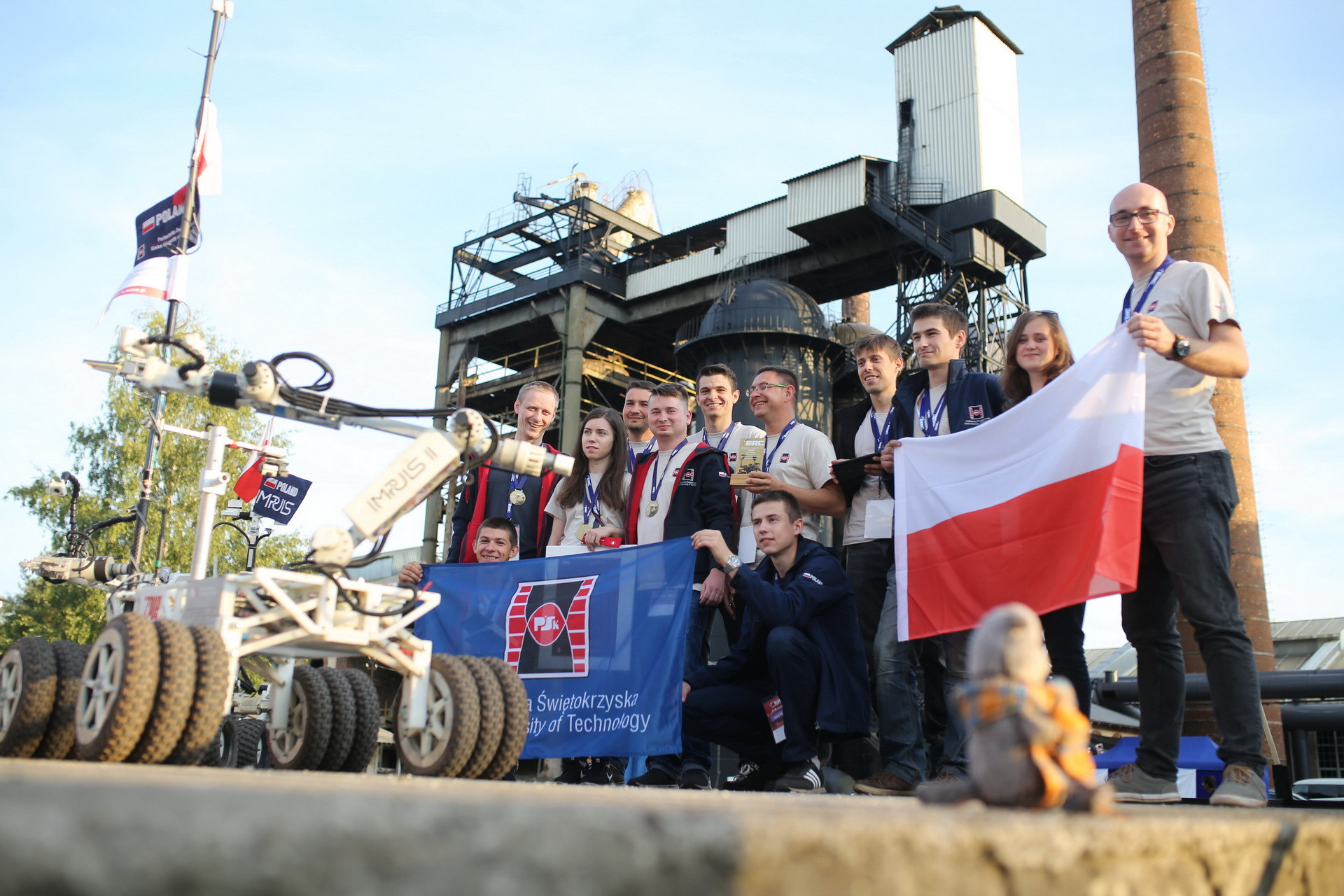 Grupa młodych ludzi z robotem i flagą Polski na tle przemysłowej budowli.