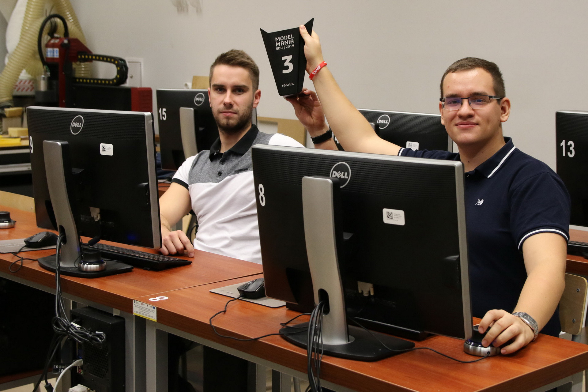 Dwóch mężczyzn siedzi przy komputerach. Jeden z nich trzyma tabliczkę z cyfrą 3. Kontekst: wydarzenie "Model Mania EDU 2019".